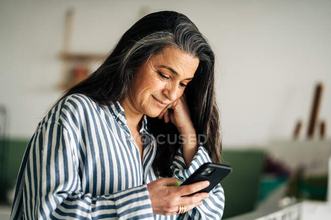 Vista lateral de la mensajería de texto femenina de mediana edad positiva en el teléfono celular mientras está sentado en la habitación con pintura creativa y caballete sobre fondo borroso - foto de stock