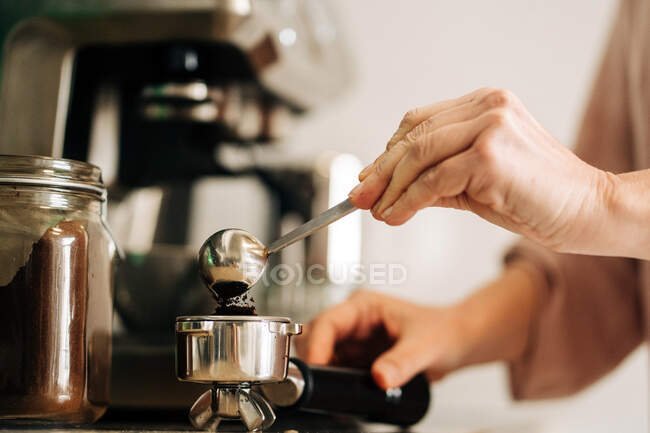 Unrecognizable female with spoon pouring ground coffee into portafilter while standing at kitchen counter with jar of coffee and coffee machine on blurred background — Stock Photo