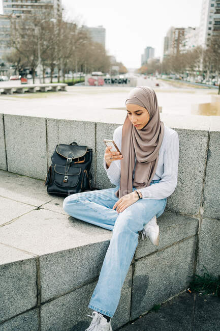 Junge ethnische Frau im Hijab sitzt auf Steinumrandung in der Stadt und sendet Nachrichten per Handy — Stockfoto