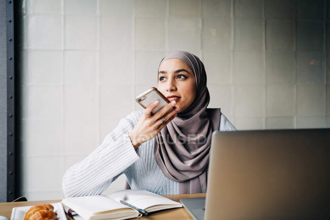 Conteúdo feminino freelancer étnico no hijab gravando mensagem de áudio no smartphone enquanto se senta à mesa no café e trabalha remotamente — Fotografia de Stock