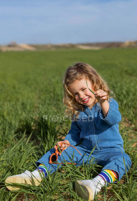 Cute happy child sitting in green field on sunny day looking at camera and playing with grass in summer — Stock Photo