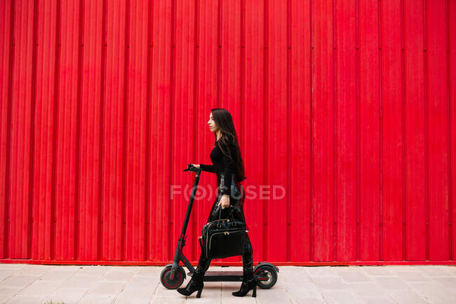 Вид збоку стильна жінка-підприємець, що йде уздовж тротуару в місті з електричним скутером на червону стіну — стокове фото