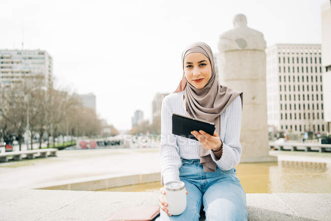 Zufriedene Muslimin mit Kopftuch sitzt mit Imbissgetränk in der Nähe des Brunnens in der Stadt und blättert in der Kamera — Stockfoto