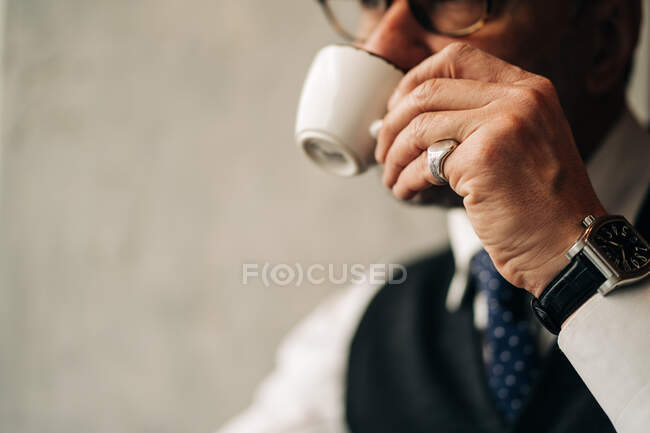 Crop empresario masculino étnico en desgaste formal y reloj de pulsera disfrutando de la bebida caliente de la taza mientras mira hacia otro lado en la cafetería - foto de stock