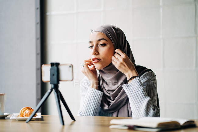 Low angle of cheerful Muslim female in headscarf shooting video on smartphone on tripod for blog while sitting at table in cafe — Stock Photo