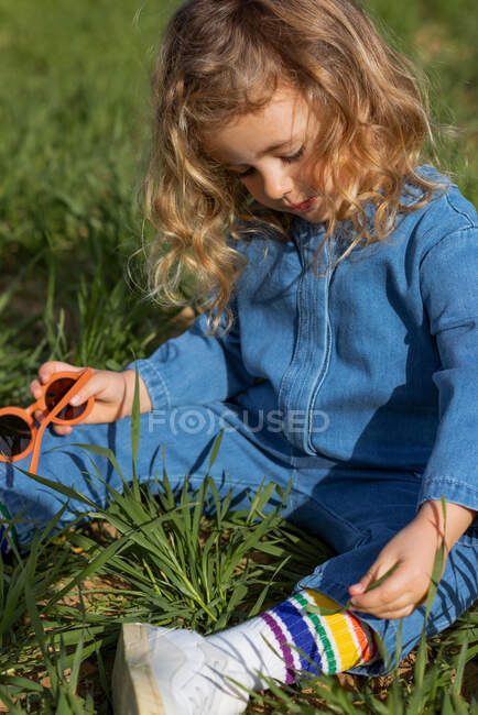 Симпатичный ребенок, сидящий на зеленом поле в солнечный день и играющий с травой летом — стоковое фото