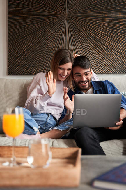 Alegre joven pareja multirracial en trajes casuales sonriendo y agitando las manos mientras se sienta en el sofá y tener una conversación de vídeo a través de la computadora portátil - foto de stock