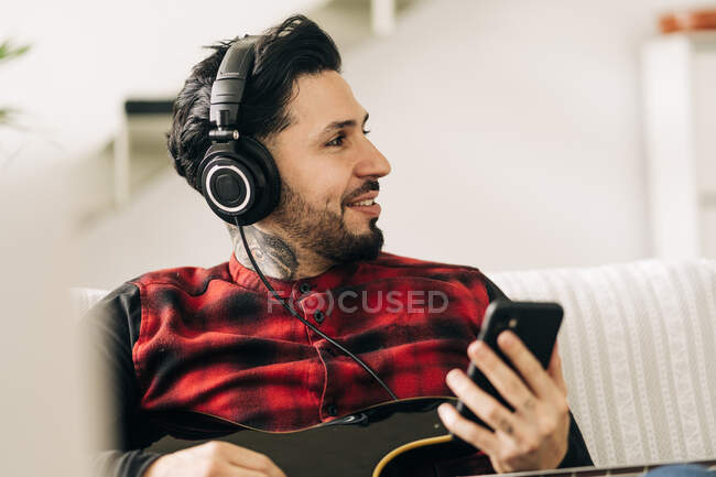 Erwachsene bärtige männliche Musiker mit Bassgitarre in Kopfhörer SMS auf Handy im Wohnzimmer — Stockfoto