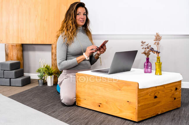 Mujer joven con pelo largo en ropa casual mensajería en el teléfono inteligente mientras trabaja de forma remota en el ordenador portátil sentado en el suelo en una pequeña mesa de madera en un apartamento minimalista - foto de stock