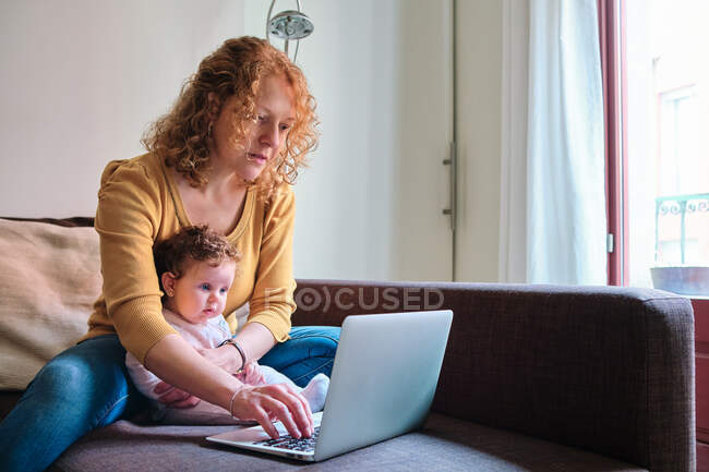 Mãe solteira freelancer sentado com bebê no sofá e netbook de navegação em casa durante o dia — Fotografia de Stock