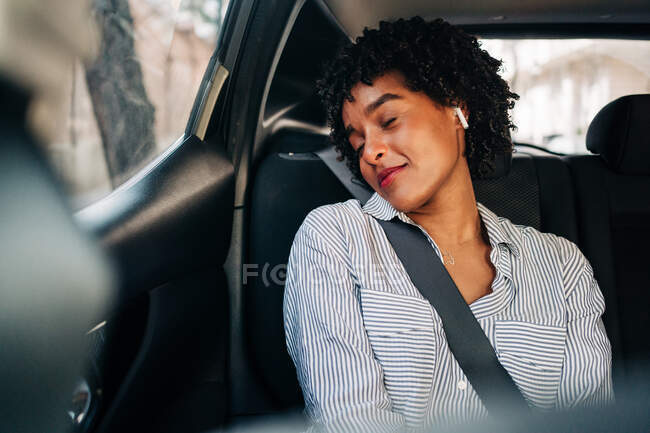Despreocupada joven afroamericana con auriculares TWS escuchando música con los ojos cerrados descansando en un automóvil moderno - foto de stock