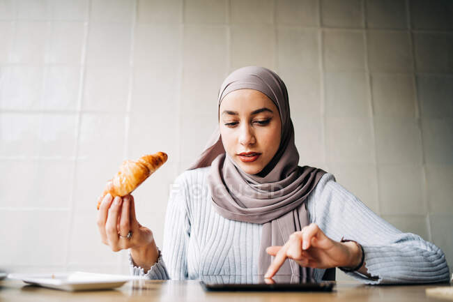 Hembra étnica en hijab navegar por Internet en la tableta mientras está sentado en la mesa con croissant y taza de café en la cafetería y disfrutar de fin de semana - foto de stock