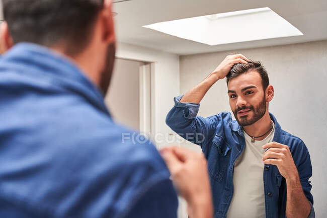 Вид сбоку сосредоточенного молодого бородатого этнического парня в стильной одежде, трогающего волосы, глядя в зеркало в современной ванной комнате — стоковое фото