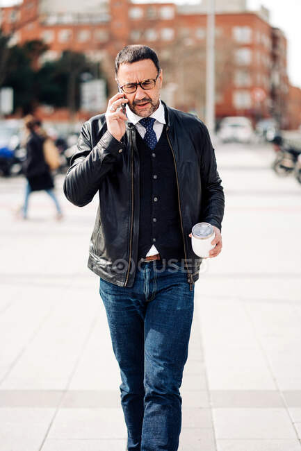 Mature hispanique mâle exécutif dans smart casual porter avec boisson chaude pour aller parler sur téléphone portable tout en marchant sur la chaussée urbaine — Photo de stock