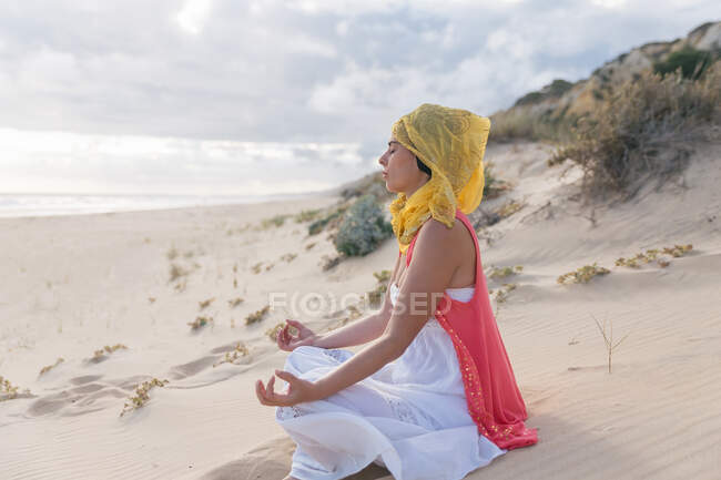 Vista lateral de una joven hembra con los ojos cerrados en sedoso pañuelo para la cabeza relajante y meditando en Padmasana en la orilla arenosa cerca del mar - foto de stock