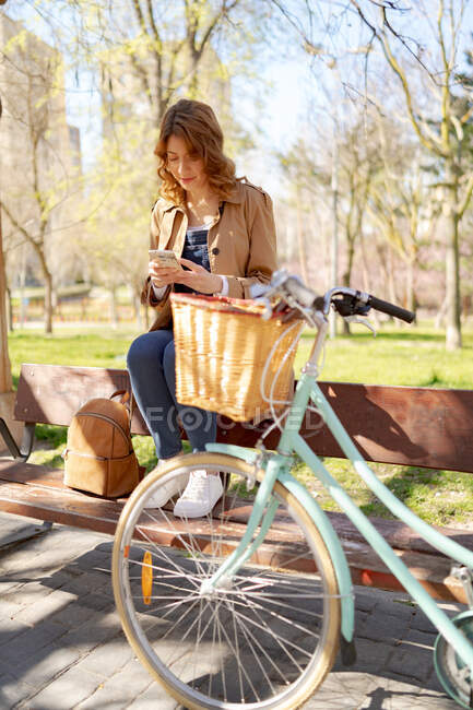 Full body of young female texting message on mobile phone near bicycle with wooden wicker basket — Stock Photo
