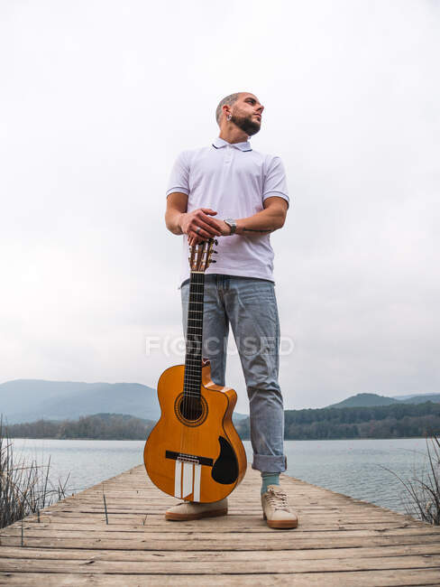 Full body of bearded guy in casual clothes standing with guitar on wooden pier near grass and river with mountains on background under cloudy gray sky in daytime — Stock Photo