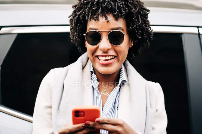 Mujer afroamericana riéndose con la boca abierta con gafas de sol elegantes usando teléfono móvil mientras está de pie cerca de un vehículo moderno - foto de stock