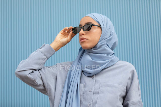 Joven mujer contemplativa étnica en pañuelo azul y gafas de sol modernas mirando hacia otro lado a la luz del día - foto de stock