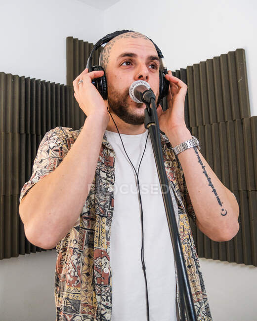 Focused man musician in stylish clothes in headphones standing and singing into microphone while recording song in studio — Stock Photo