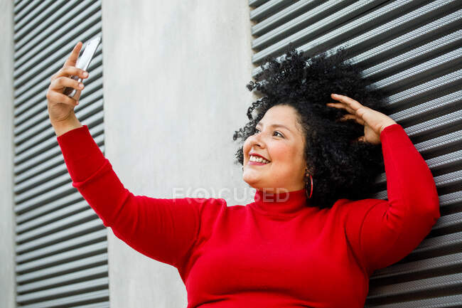 Vista lateral da fêmea sorridente gorda em desgaste brilhante fazendo um auto-retrato enquanto se inclina na parede com nervuras na cidade — Fotografia de Stock