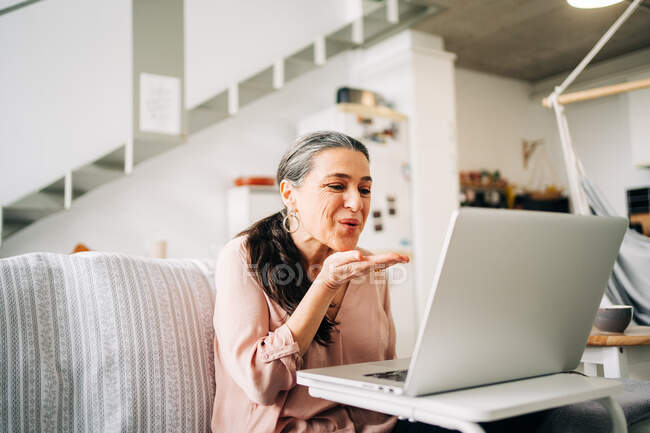 Positive middle aged female sitting on couch and sending air kiss while having video chat on netbook in modern apartment — Stock Photo
