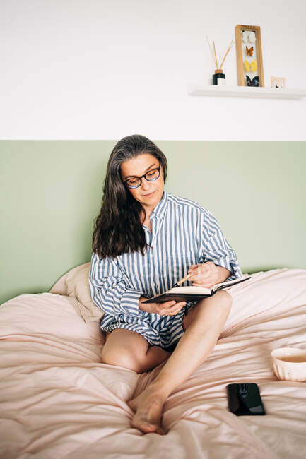 Middle aged cheerful female in striped shirt smiling and writing notebook on bed with smartphone — Stock Photo