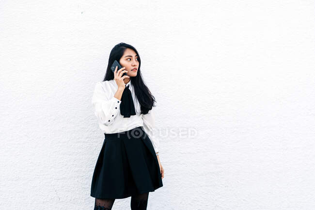 Adolescent ethnique asiatique femelle en tenue formelle debout près du mur blanc parlant sur le téléphone mobile — Photo de stock