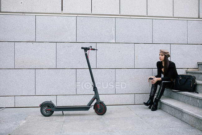 Side view of styles female entrepreneur in beret hat sitting on staircase in city near modern electric scooter — Stock Photo