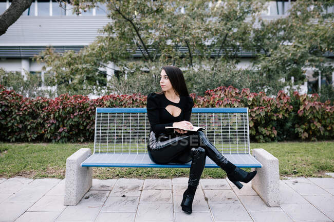 Stylish female entrepreneur sitting on bench and taking notes in organizer while working in urban park — Stock Photo