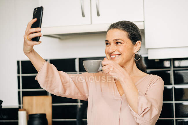 Зачарована жінка середнього віку з чашкою гарячого напою, що приймає власний портрет, стоячи на кухні біля білих шаф вдома — стокове фото
