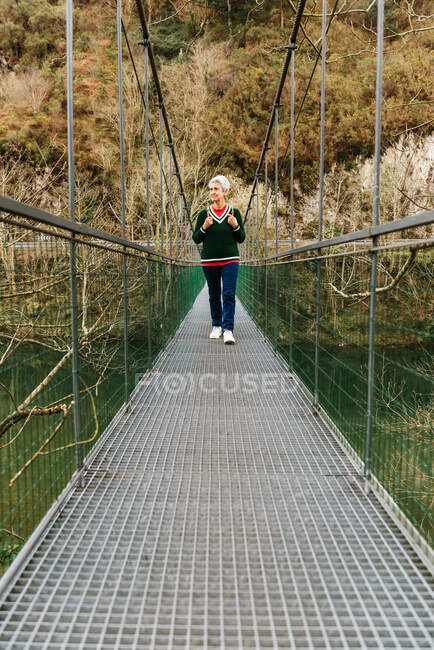 Elderly female hiker in casual clothes strolling on suspension bridge while looking away in daytime — Stock Photo