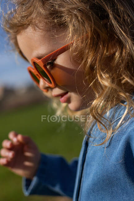 Вид сбоку симпатичной счастливой маленькой девочки в трикотажных одеждах и солнцезащитных очках, стоящей и расслабляющейся на расстегнутом пуховике — стоковое фото