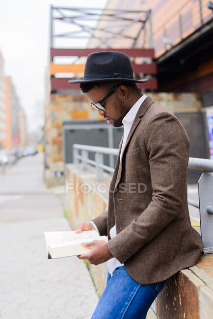 Vista lateral do homem afro-americano em chapéu elegante e jaqueta de pé na rua da cidade e lendo livro interessante — Fotografia de Stock