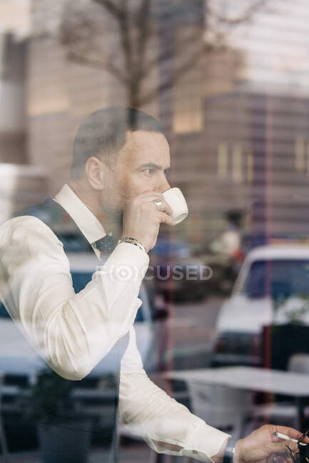 Durch Glaswand Seitenansicht eines älteren hispanischen männlichen Unternehmers in formalem Outfit, der Heißgetränk genießt, während er sich auf das Café freut — Stockfoto