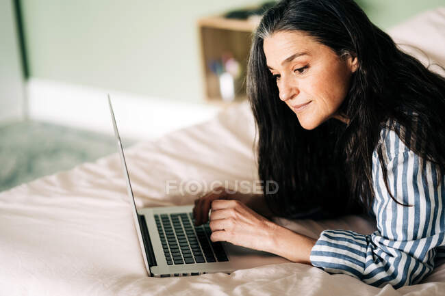 Vue latérale de la femme hispanique mature autonome focalisée avec de longs cheveux foncés dans des vêtements décontractés couchés sur le lit et tapant sur un ordinateur portable pendant le travail en ligne à la maison — Photo de stock