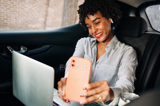 Cheerful African American female smiling and taking selfie with mobile phone in modern automobile — Stock Photo