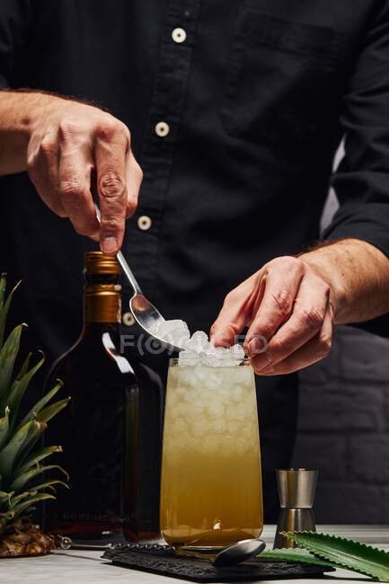Crop anonimo barman mettendo cubetti di ghiaccio da cucchiaio in vetro con cocktail su sottobicchiere vicino foglie verdi e ananas vicino bottiglia sul bancone vicino muro di mattoni — Foto stock