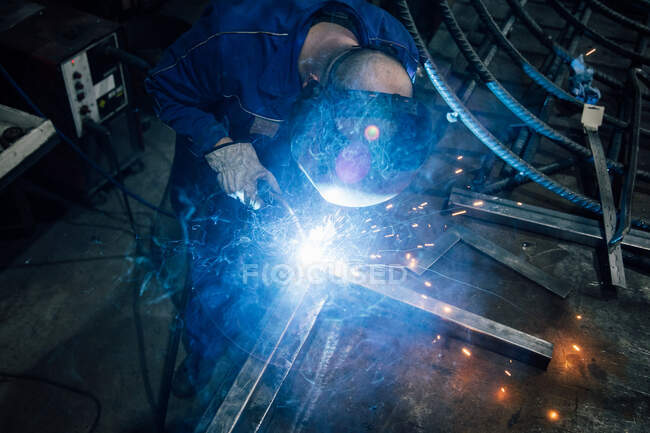 Crop faceless worker in gloves and uniform welding metal details on table near constructions in factory — Stock Photo