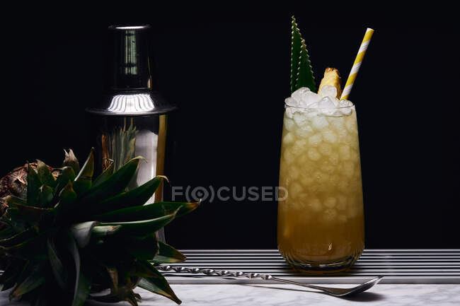 Желтый коктейль в стекле, украшенный кусочком ананаса и зелеными листьями с бумажной соломинкой возле барной ложки — стоковое фото