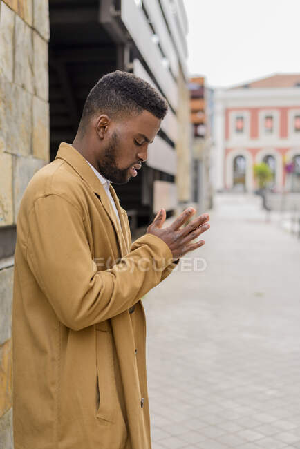 Side view African American male wearing trendy coat standing near stone wall of building in city and putting hands together — Stock Photo
