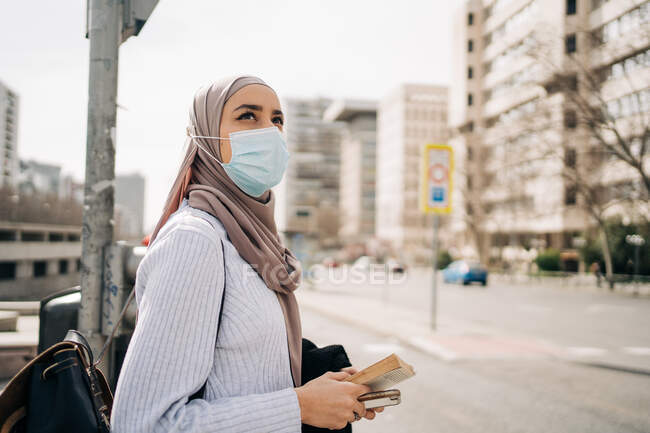Side view of ethnic female wearing headscarf and protective mask standing on street in city on sunny day and looking away — Stock Photo