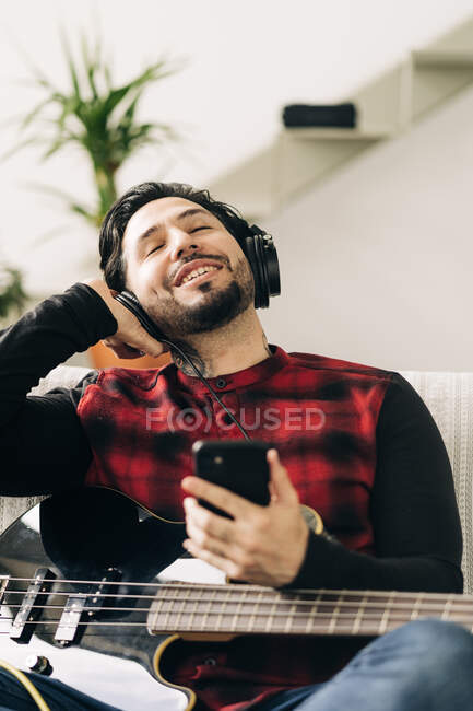 Musicien homme barbu adulte avec guitare basse dans casque messagerie texte sur téléphone portable dans le salon — Photo de stock