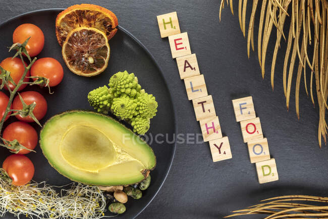 Top view of decorative inscription near plate with fresh half of avocado and bundle of cherry tomatoes near red orange slices — Stock Photo