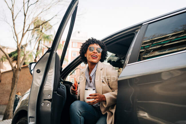 Happy African American female in fashionable sunglasses with thermal mug and using smartphone while smiling and leaving silver prestige automobile — Stock Photo