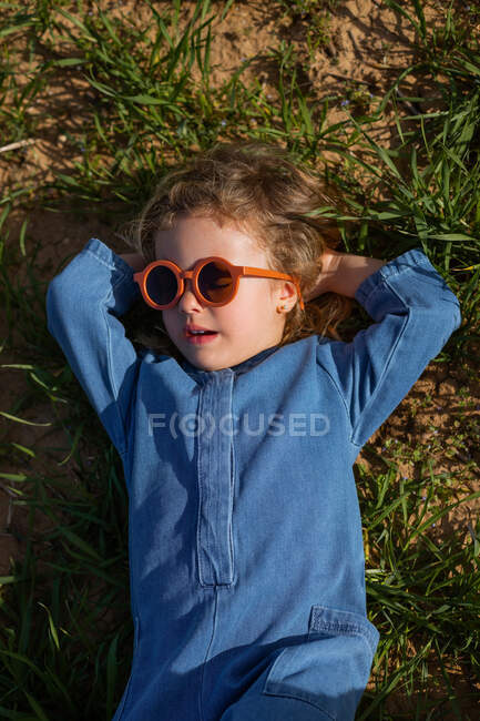 From above girl in trendy clothes and sunglasses holding hands behind head and relaxing on grassy lawn — Stock Photo