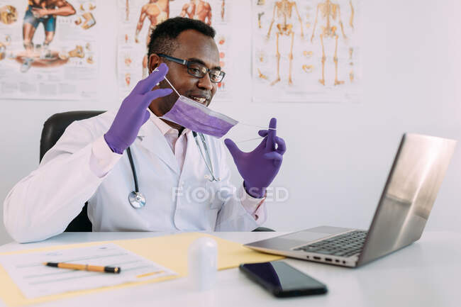 Médico afroamericano en anteojos y mascarilla facial trabajando con paciente en línea en netbook mientras escribe en archivo de paciente en la mesa en el hospital - foto de stock