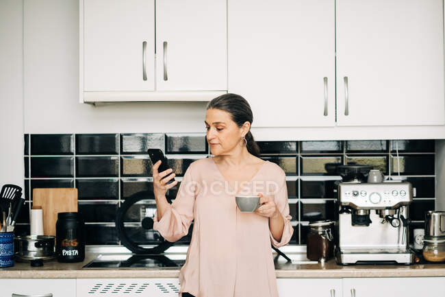 Mulher de meia-idade encantada com xícara de bebida quente navegando no smartphone na cozinha perto de armários brancos em casa — Fotografia de Stock