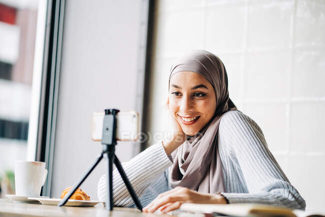 Bajo ángulo de alegre mujer musulmana en el pañuelo para la cabeza de vídeo en el teléfono inteligente en el trípode para el blog mientras está sentado en la mesa en la cafetería - foto de stock