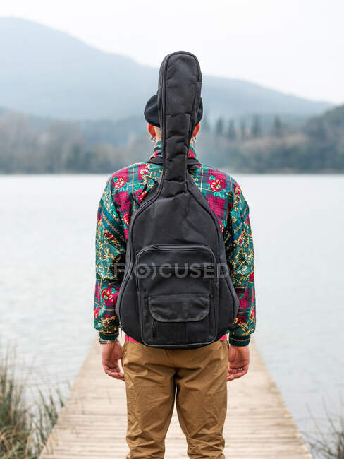 Back view of thoughtful male in stylish clothes standing with guitar in case on wooden pier near calm river against mountain in cloudy day — Stock Photo
